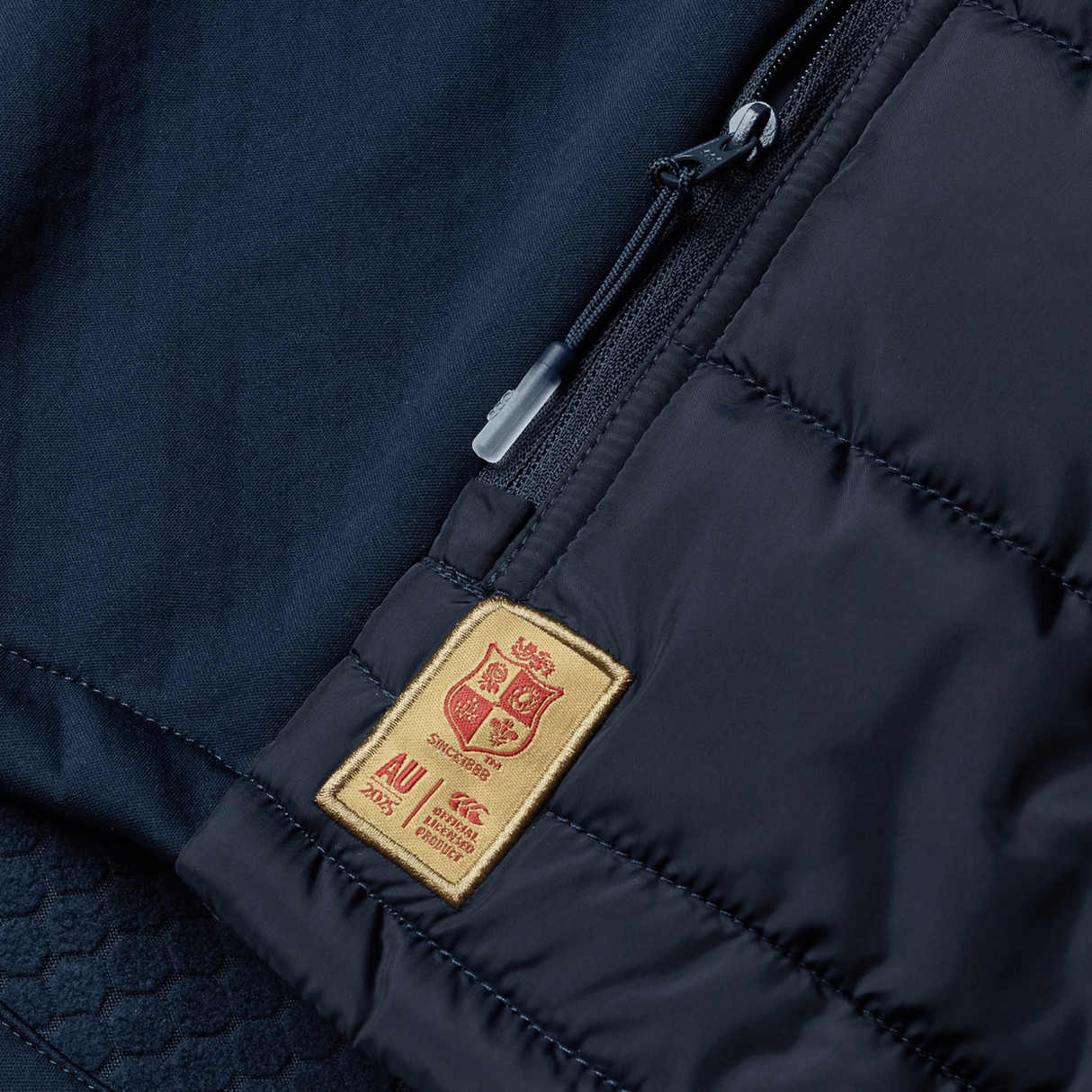 A close-up of the British & Irish Lions 2025 Microlight Gilet by Canterbury showcases its black thermoreg fabric with a textured surface, accented by a small beige patch featuring red embroidered letters. The piece also includes a partially open zippered pocket, blending sleek design with the sustainable appeal of recycled polyester.