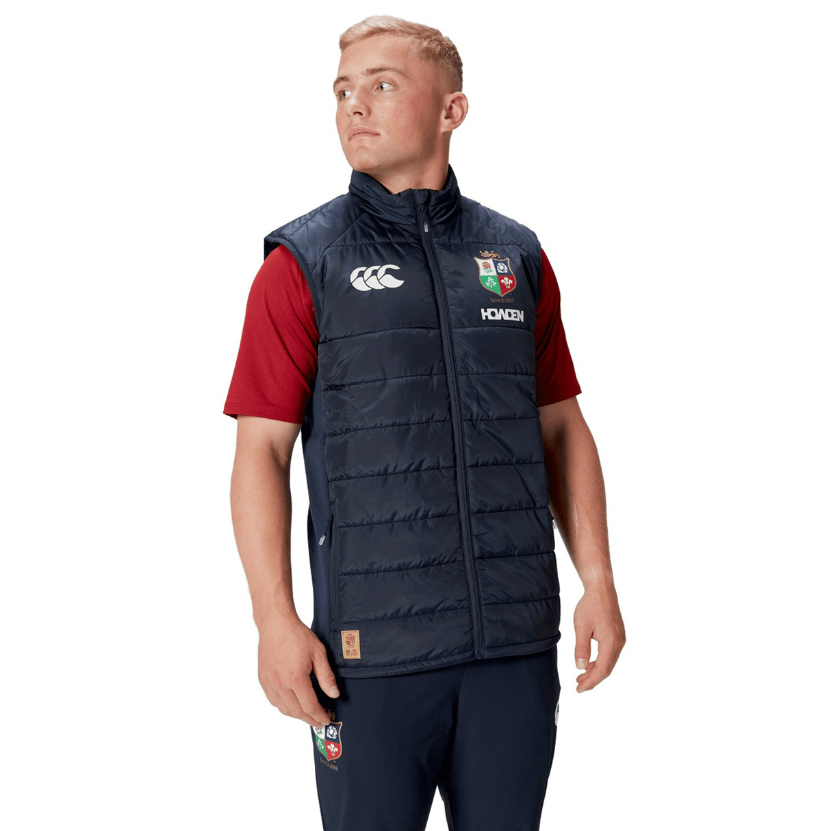 A person wearing a British & Irish Lions 2025 Microlight Gilet by Canterbury in navy blue, made from recycled polyester, over a red shirt and navy pants, looks to the side.