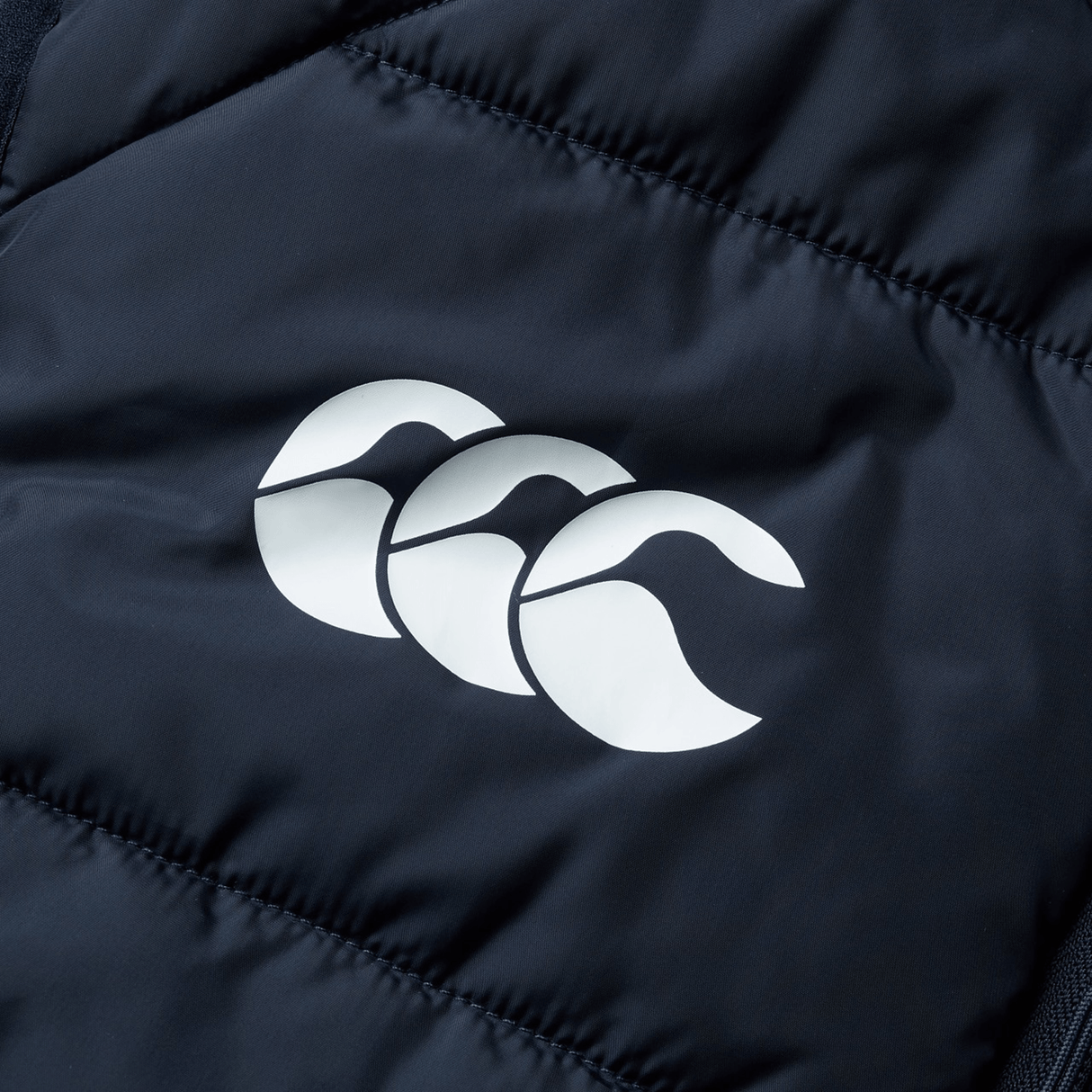 Close-up of the British & Irish Lions 2025 Microlight Gilet by Canterbury, featuring a dark fabric with a white logo of three overlapping stylized wave shapes, made from recycled polyester.