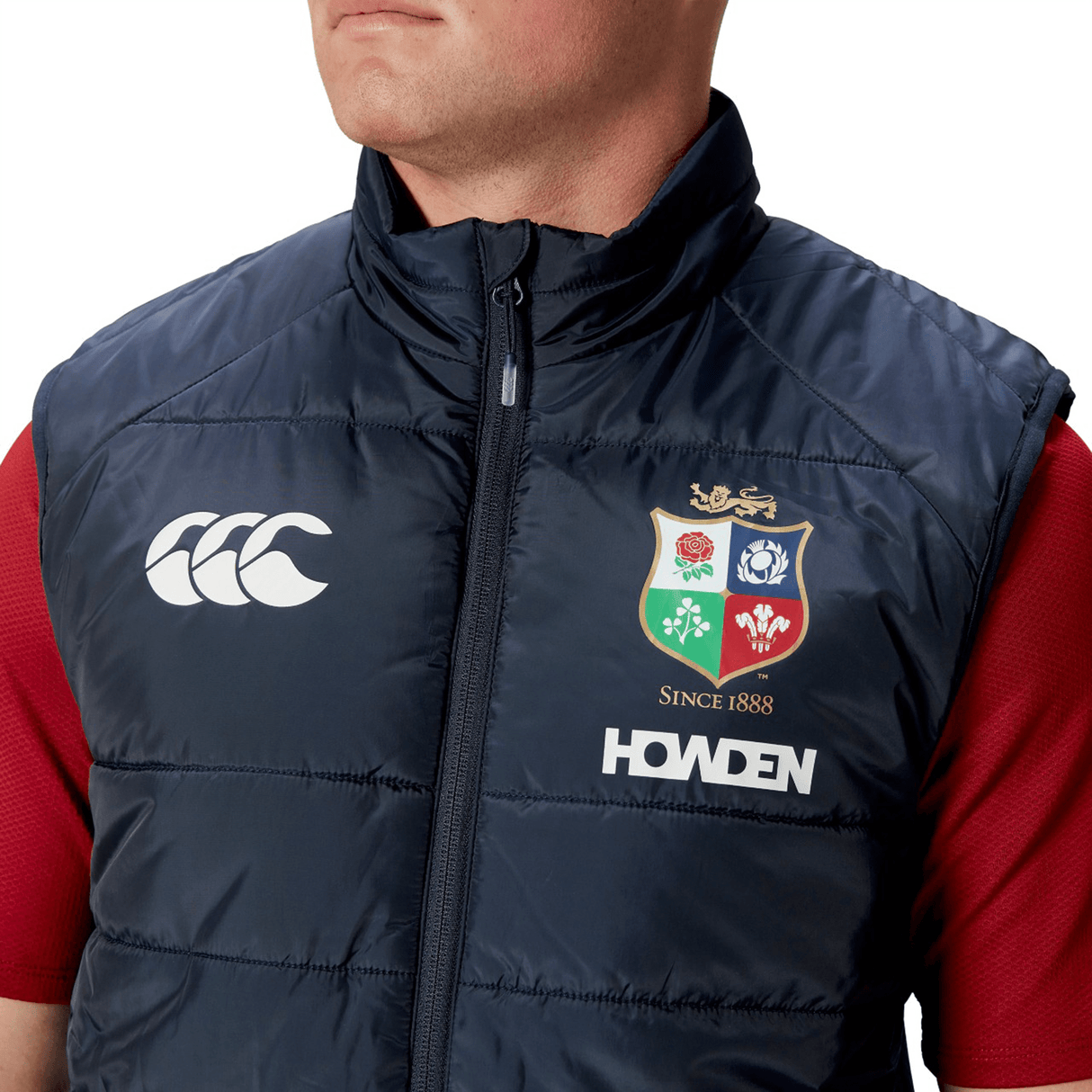 Dressed in a British & Irish Lions 2025 Microlight Gilet by Canterbury, this individual showcases a navy blue vest adorned with the iconic Canterbury logo, the British and Irish Lions crest, and "HOWDEN" lettering. Made from recycled polyester with thermoreg technology, the gilet is fashionably paired over a red shirt.