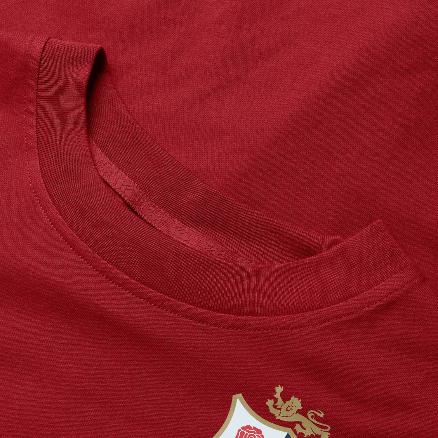 Close-up of the Women's British & Irish Lions 25 Cotton Jersey Tee by Canterbury. This red T-shirt is crafted from organic cotton and features a distinctive shield emblem adorned with a white rose and gold lion in the lower corner, making it an ideal choice for any rugby supporter. The detailed design proudly highlights both the neckline and fabric texture.