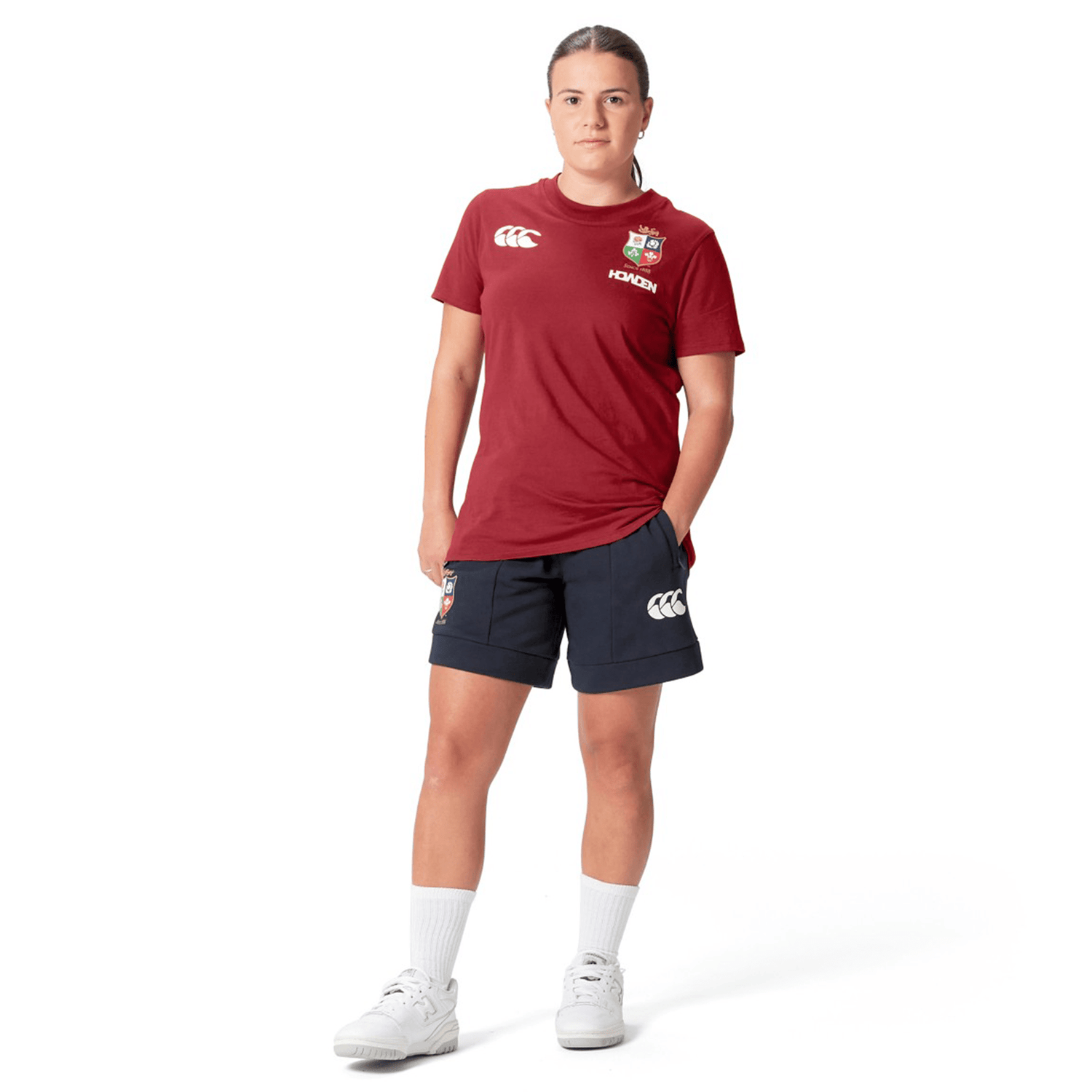 A person stands wearing a Women's British & Irish Lions 25 Cotton Jersey Tee by Canterbury in red, along with navy shorts and white sneakers, against a white background.