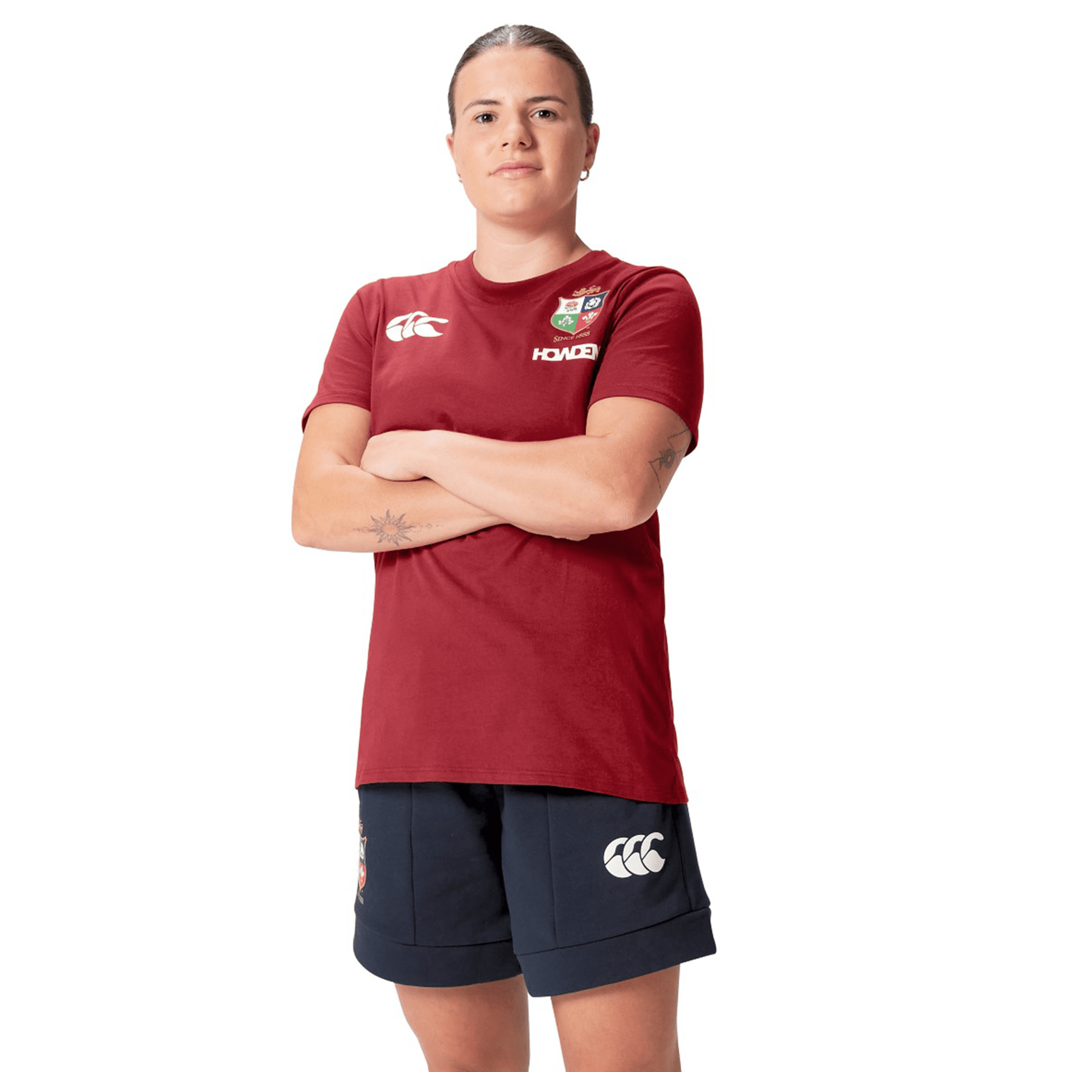 A rugby supporter in the Women's British & Irish Lions 25 Cotton Jersey Tee by Canterbury, paired with navy shorts, stands with arms crossed. The outfit is made from organic cotton.