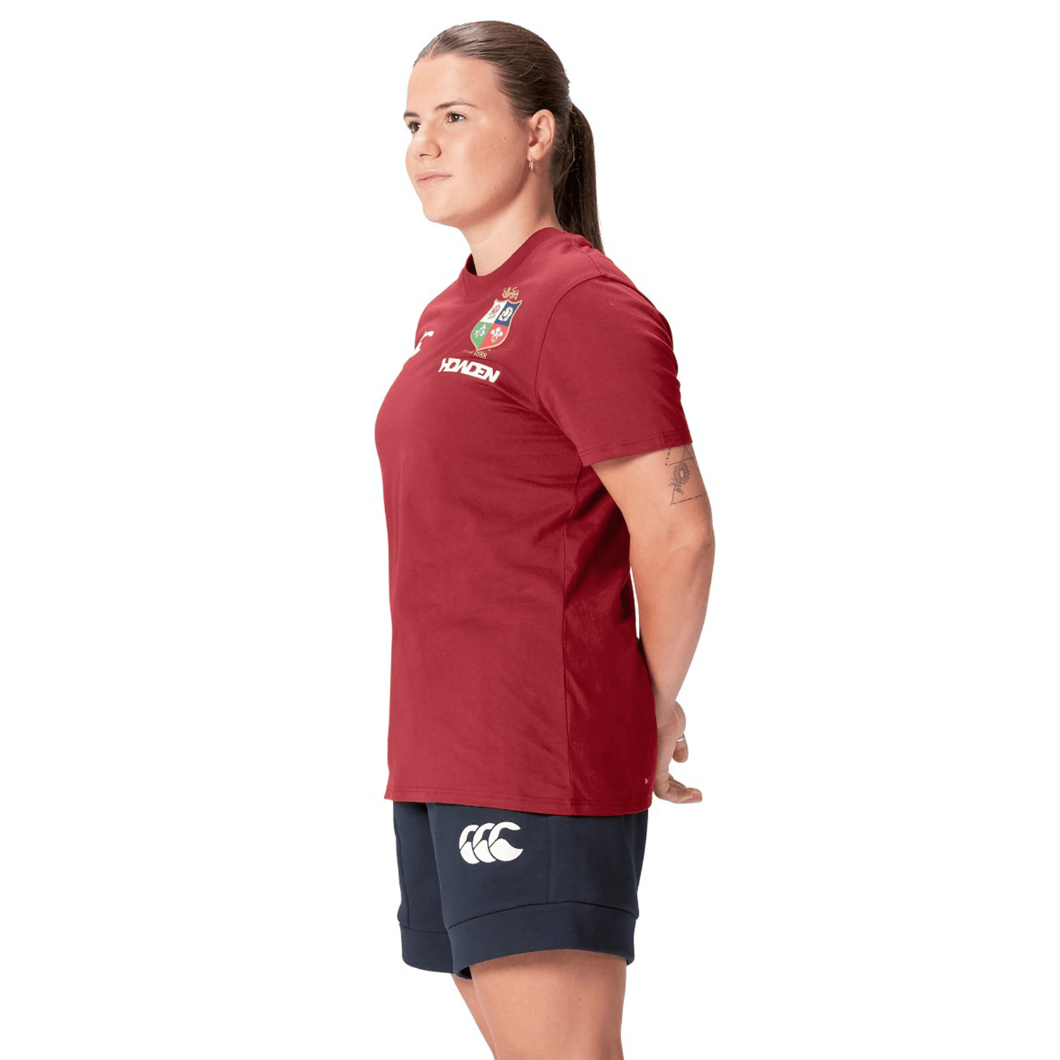 A rugby fan wearing the Women's British & Irish Lions 25 Cotton Jersey Tee by Canterbury and navy shorts stands with their hands behind their back, facing left.