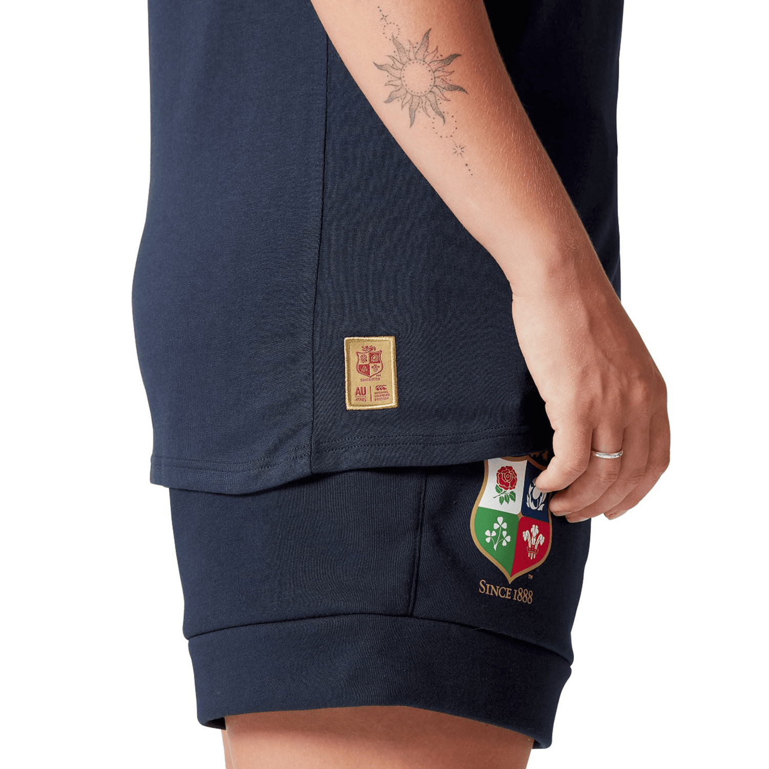 A close-up of a person wearing the Women's British & Irish Lions 25 Cotton Jersey Tee by Canterbury in navy, featuring a sun tattoo on the arm and a patch with four emblems on the leg, captures the essence of every rugby supporter. Made from organic cotton, this jersey embodies sustainable style while celebrating the spirit of the British & Irish Lions.