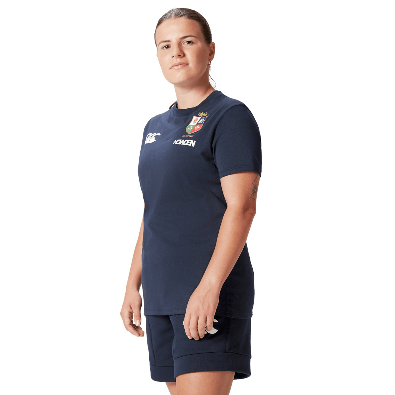 A rugby fan in a navy blue *Women's British & Irish Lions 25 Cotton Jersey Tee by Canterbury* stands against a white background, looking confidently at the camera.