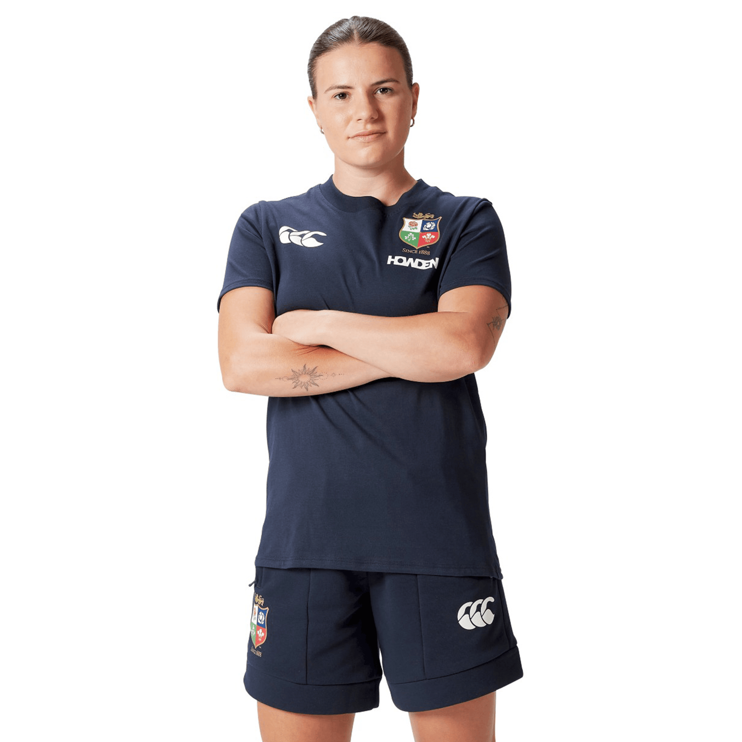 A rugby supporter in navy sportswear stands confidently with arms crossed, showcasing a Women's British & Irish Lions 25 Cotton Jersey Tee by Canterbury and shorts featuring logos. This attire, made from organic cotton, proudly represents the spirit of the British & Irish Lions.