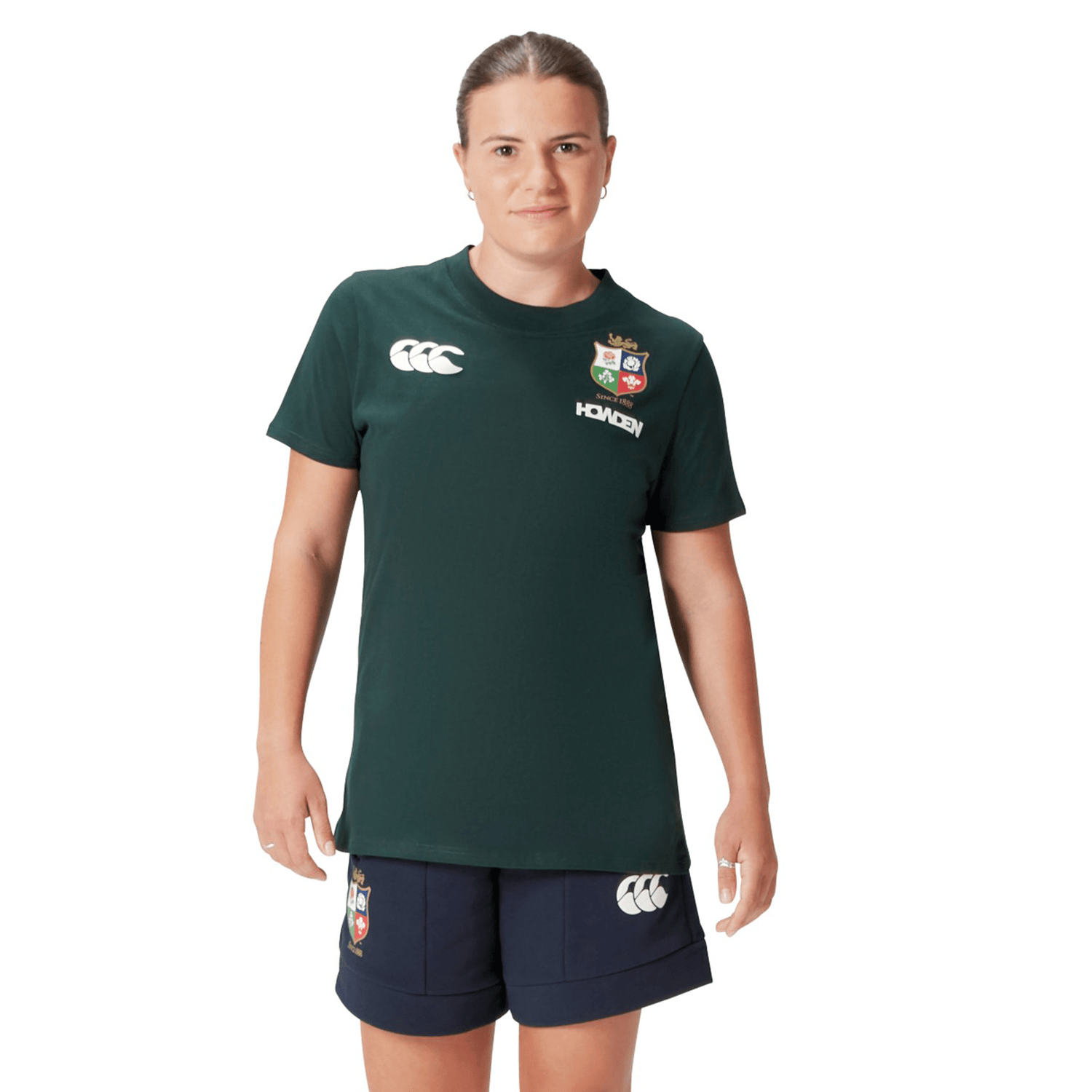 A rugby supporter wearing the Women's British & Irish Lions 25 Cotton Jersey Tee by Canterbury, featuring a dark green color and made from organic cotton, paired with navy shorts adorned with logos, stands against a white background.