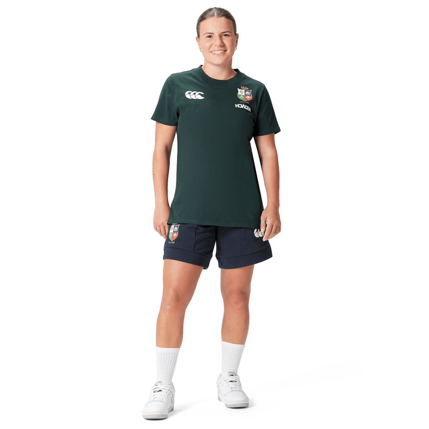 A person dressed in the Women's British & Irish Lions 25 Cotton Jersey Tee by Canterbury, complemented by navy shorts, white socks, and sneakers, stands against a plain white background.
