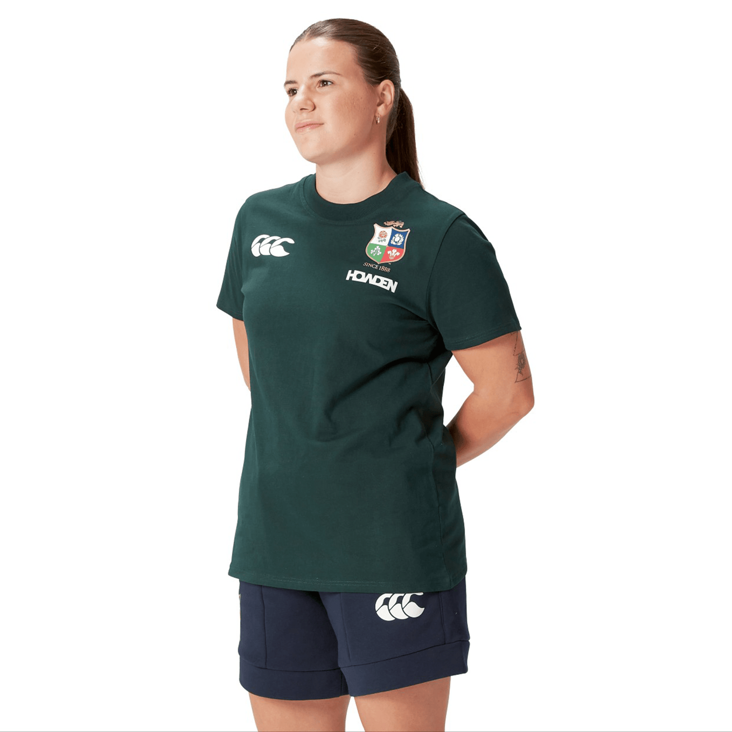 A rugby supporter proudly stands in a Women's British & Irish Lions 25 Cotton Jersey Tee by Canterbury, with navy shorts, hands clasped behind their back, glancing to the side.
