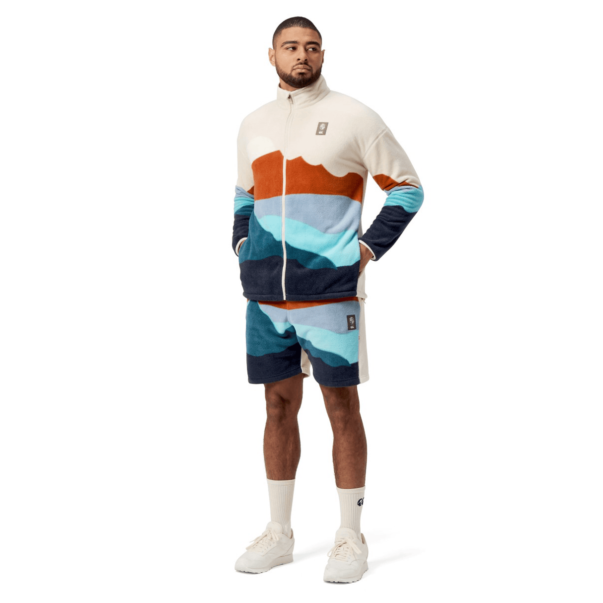 Dressed in a Canterbury British & Irish Lions 2025 Outback Full-Zip Fleece Jacket, the man strikes a pose with hands tucked into his pockets, paired with shorts featuring a bold mountain pattern. He finishes off his outfit with white socks and sneakers, capturing an adventurous spirit that echoes the essence of the British & Irish Lions.