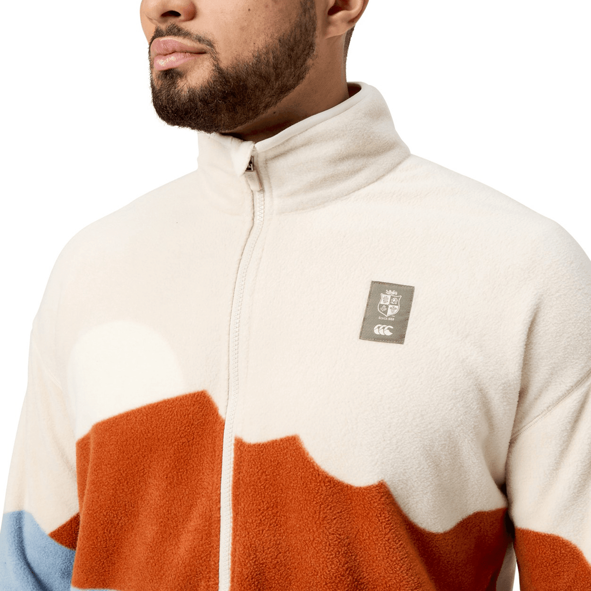 Someone is wearing a cream and rust-colored British & Irish Lions 2025 Outback Full-Zip Fleece Jacket by Canterbury, featuring a high collar and prominently displaying the Canterbury crest logo on the chest.