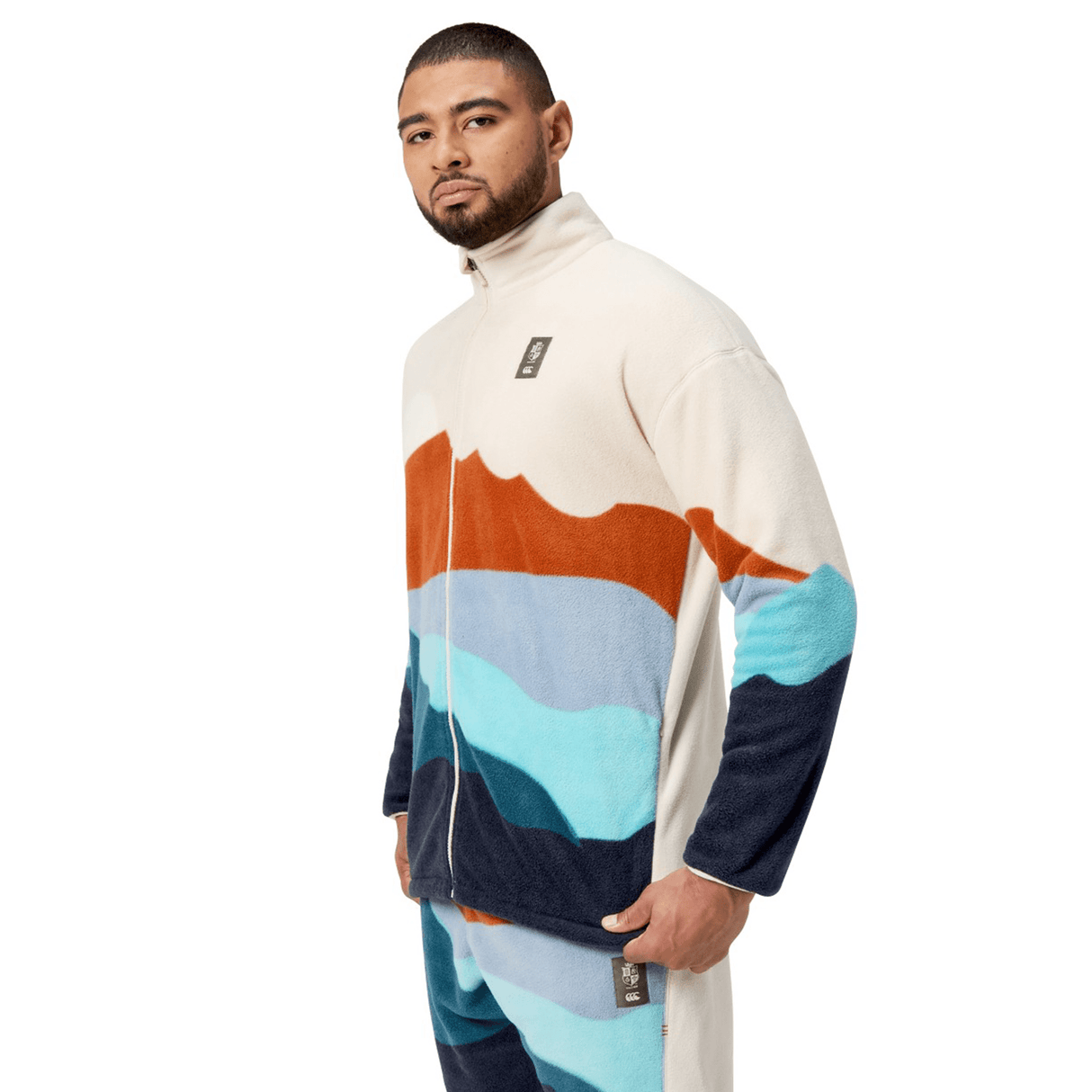 A person standing and looking forward, wearing the British & Irish Lions 2025 Outback Full-Zip Fleece Jacket by Canterbury, which features a landscape design with orange, light blue, and navy hues.