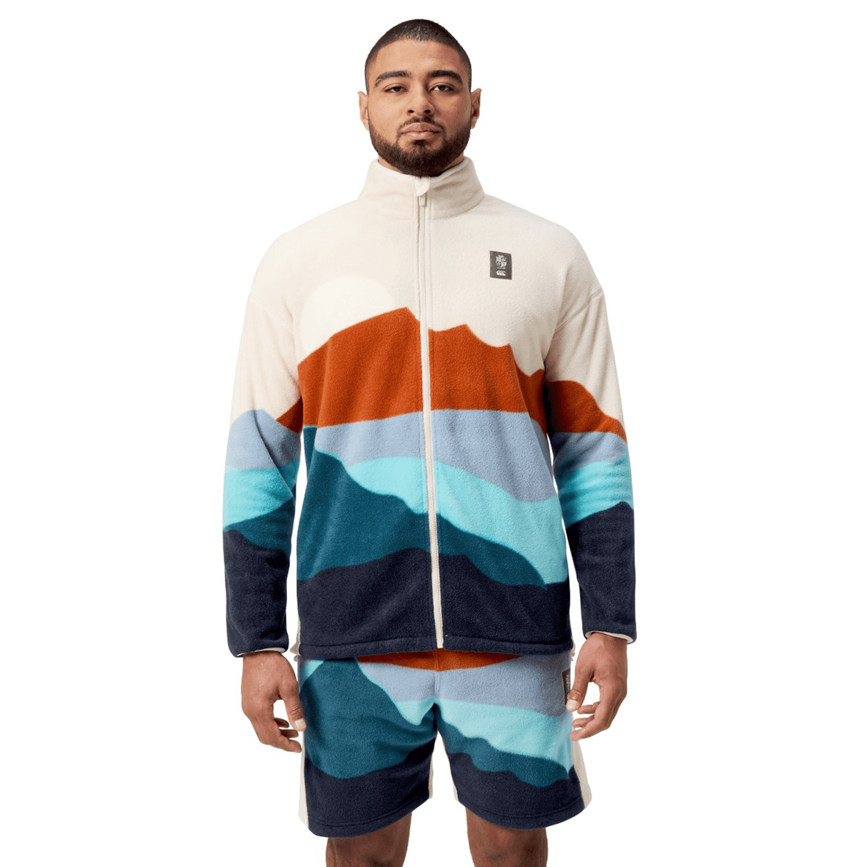 A person wears a British & Irish Lions 2025 Outback Full-Zip Fleece Jacket by Canterbury, featuring mountain-like patterns and vibrant colors, paired with shorts against a plain white background.