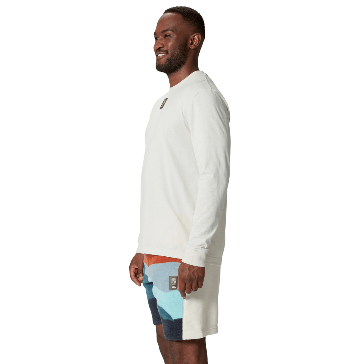 A person stands sideways wearing a British & Irish Lions 25 Outback Long Sleeve Tee by Canterbury and multicolored shorts, smiling against a plain white background.