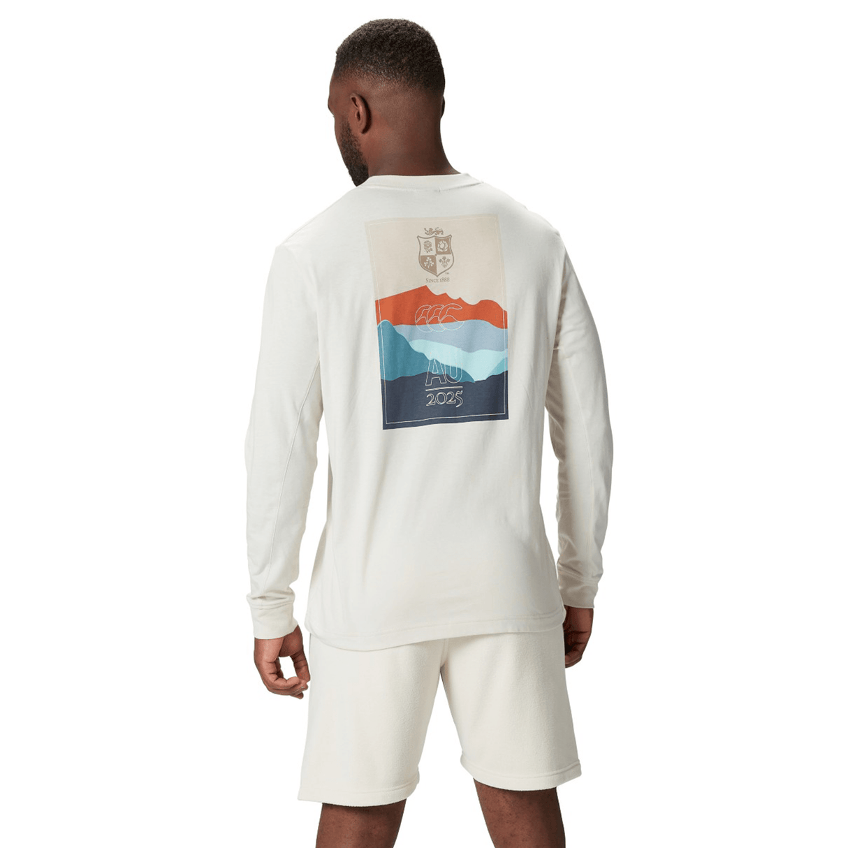 A man in a Canterbury British & Irish Lions 25 Outback Long Sleeve Tee, adorned with a multicolored graphic, paired with white shorts, stands facing away.