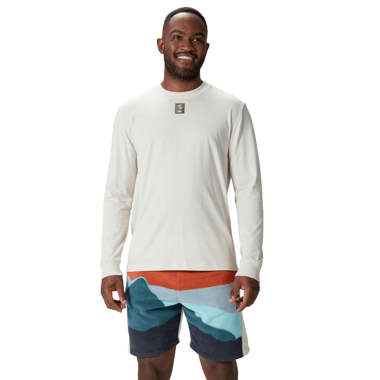 A man wearing a British & Irish Lions 25 Outback Long Sleeve Tee by Canterbury and colorful shorts stands smiling against a white background, evoking the vibrant spirit of the British & Irish Lions.