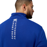 A man in a Canterbury Sport Dept 1/4 Zip Pullover stands with his back to the camera, wearing a blue jacket adorned with "Canterbury Sport Dept" in white text and featuring rib-knit cuffs for comfort and style.