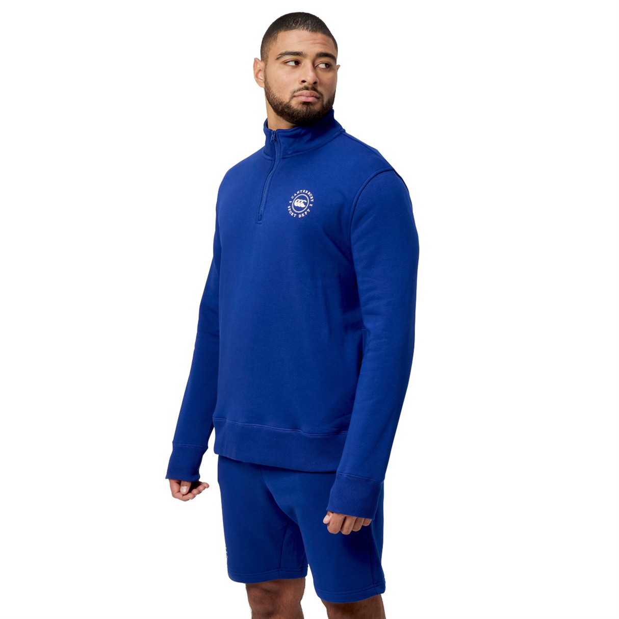 A person wearing a blue Canterbury Sport Dept 1/4 Zip Pullover with rib-knit cuffs and shorts, gazing to the side on a white background.