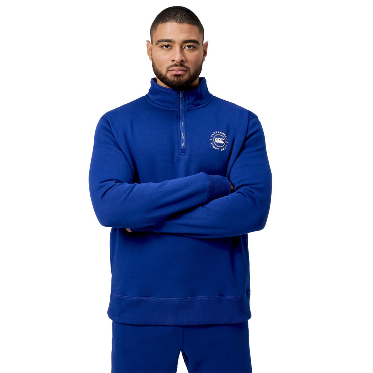 A man in a blue Canterbury Sport Dept 1/4 Zip Pullover, featuring the Canterbury logo on the chest, stands confidently with arms crossed against a white background.