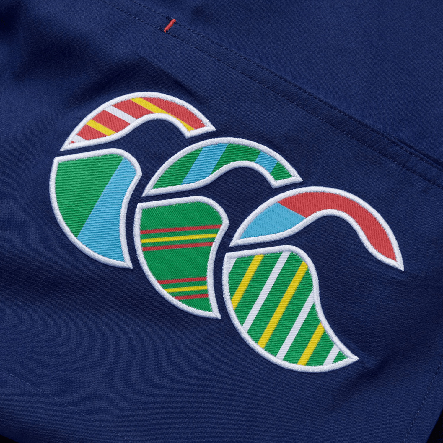 Close-up of the Canterbury Uglies 5" Tactic Short in blue with a multicolored logo showing red, green, yellow, and white stripes. This stylish design complements its deep pockets and elastic waist, offering a seamless blend of comfort and style.