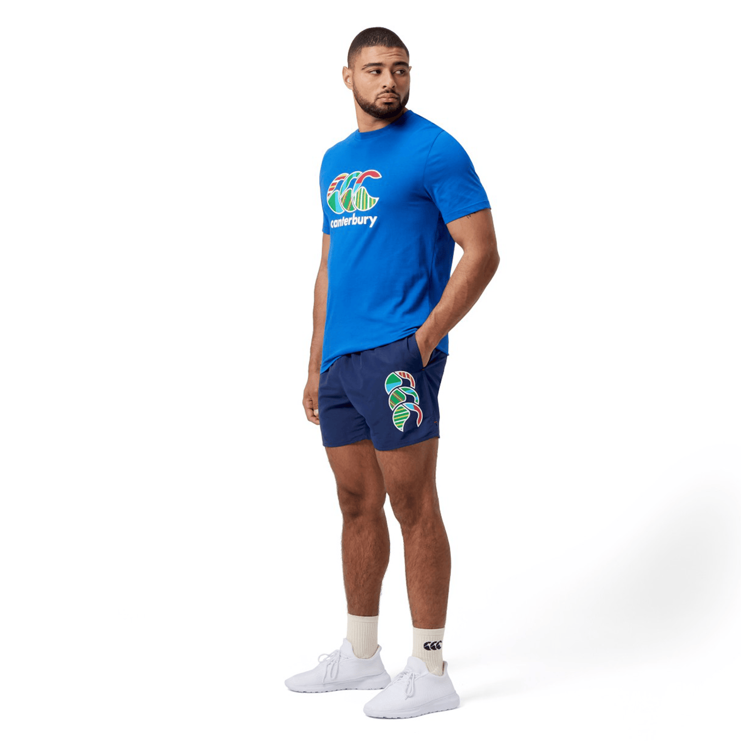 A man in a blue T-shirt pairs his Canterbury Uglies 5" Tactic Short, featuring an elastic waist and deep pockets with a colorful logo, with white sneakers and socks. He stands confidently against a white background.