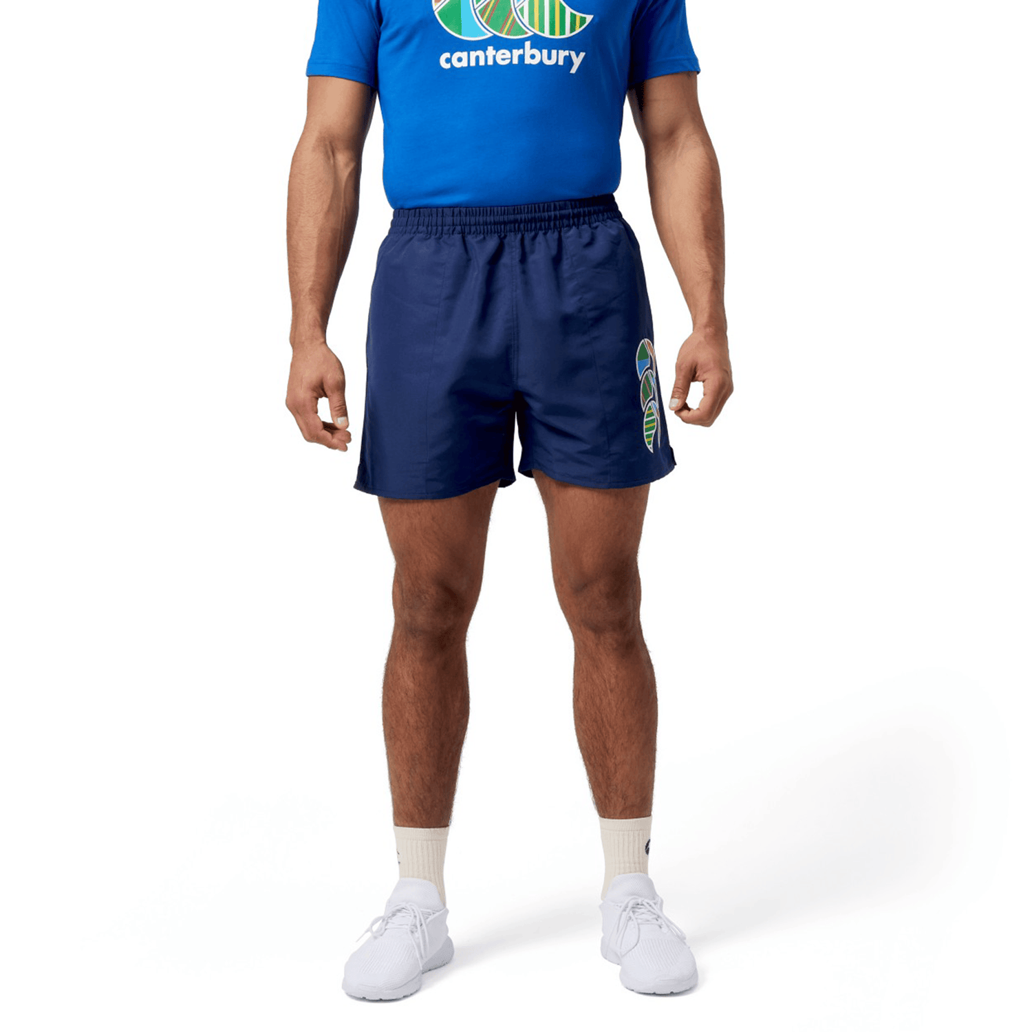 A man in a blue tee and Canterbury Uglies 5" Tactic Shorts, navy with logo, elastic waist, and deep pockets, stands against a white background.