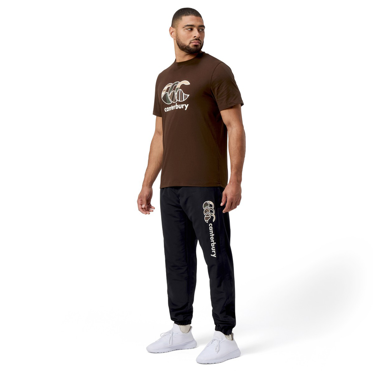 A man in a brown Canterbury t-shirt, black Canterbury Uglies Cuffed Stadium Pant joggers, and white sneakers stands against a plain white background, facing slightly to the left.