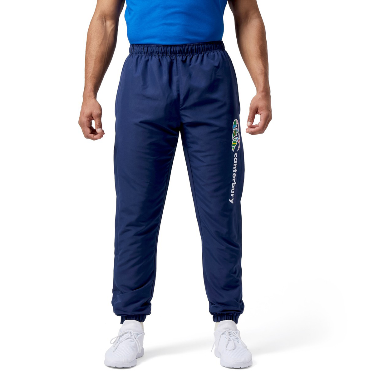 A man stands against a white background in Canterbury Uglies Cuffed Stadium Pant, paired with a blue shirt and white sneakers. The navy rugby-style pants add a distinctive touch reminiscent of the Canterbury Uglies collection.
