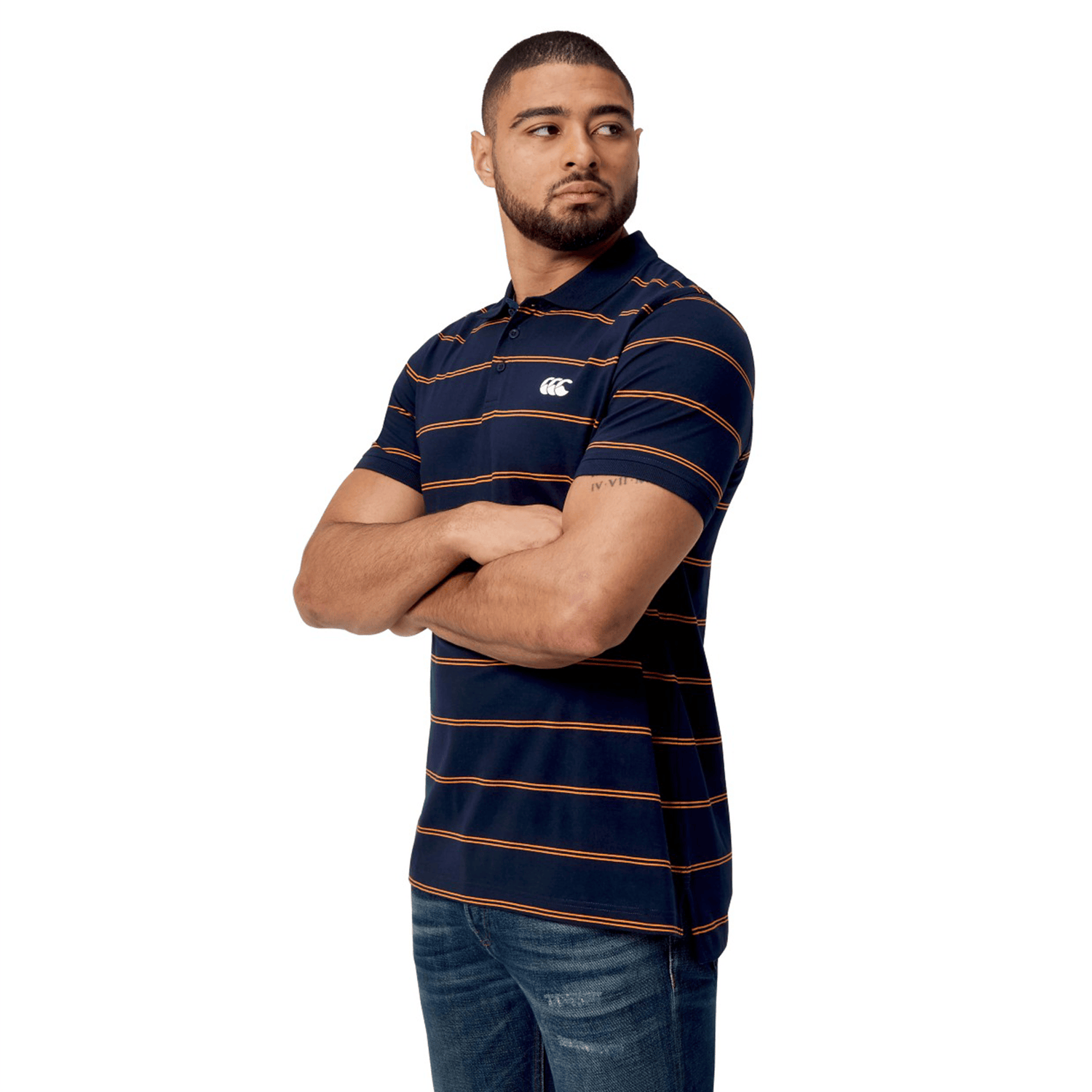 A man stands against a white background, wearing the Canterbury Fundamental Yarn-Dye Stripe Polo, featuring navy and orange stripes, paired with casual jeans. Arms confidently crossed in this stylish attire.