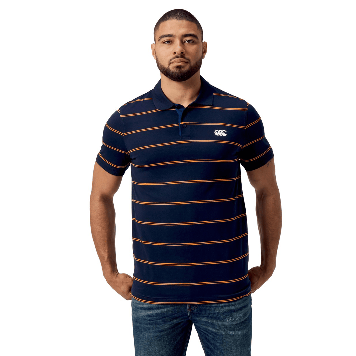 Someone wearing a Canterbury Fundamental Yarn-Dye Stripe Polo, with navy and yarn-dye stripes, and jeans stands with hands in pockets against a white background.