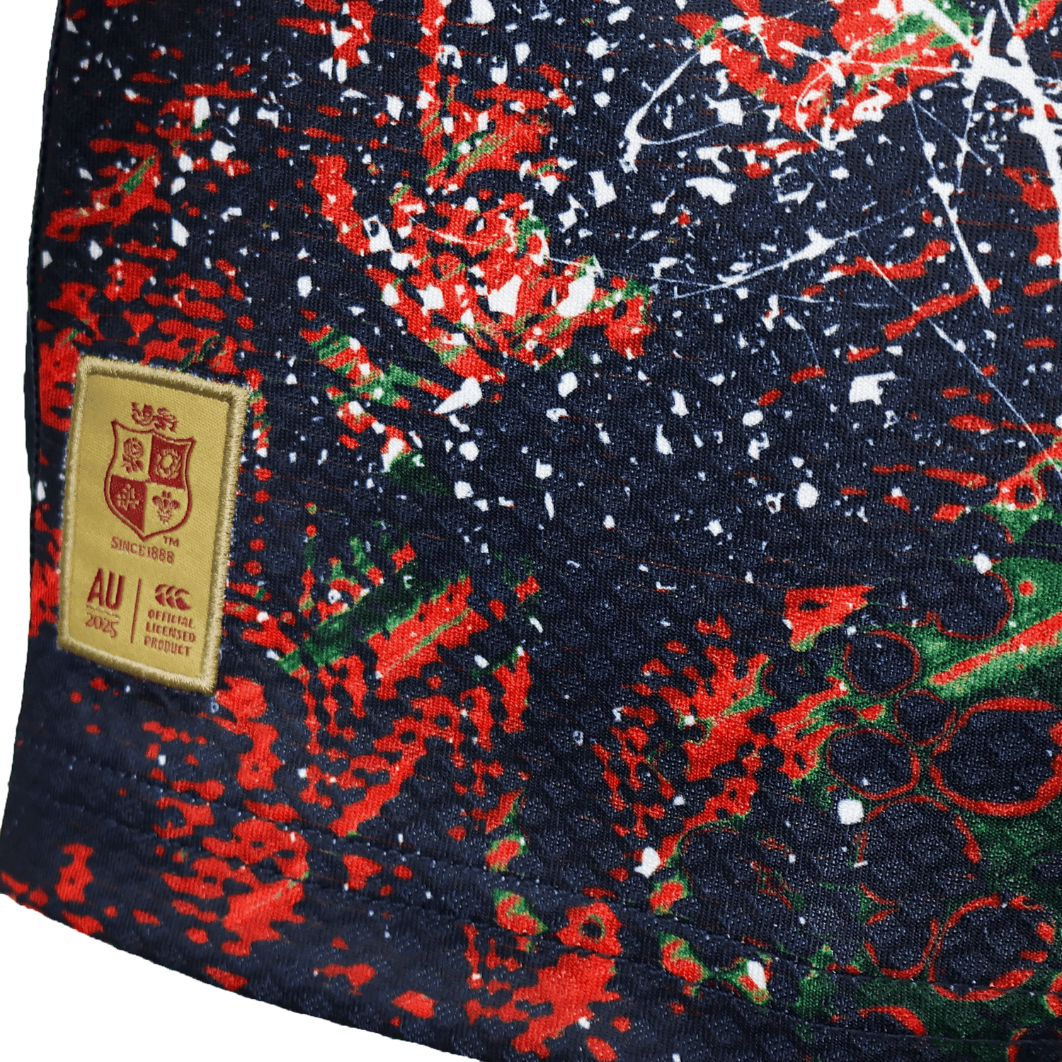 Close-up of the Youth British & Irish Lions 2025 Warm Up Jersey by Canterbury, showcasing moisture-wicking fabric featuring a vibrant abstract design in red, green, and white on a dark background, with a beige patch resembling the UK Lions crest and "AU" and "2003" text.