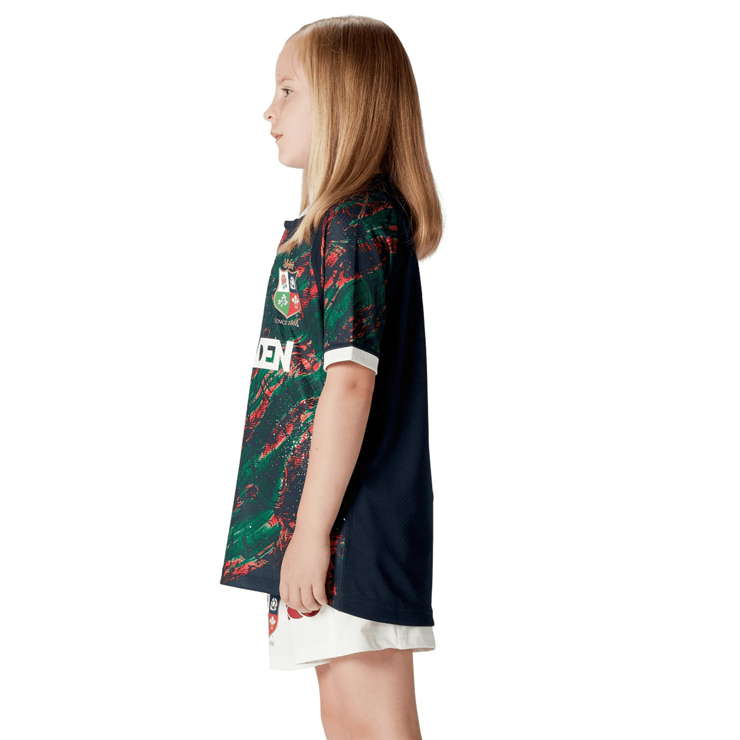 A young girl with long hair stands in profile, proudly wearing the Youth British & Irish Lions 2025 Warm Up Jersey by Canterbury, crafted from moisture-wicking fabric, paired with crisp white shorts.