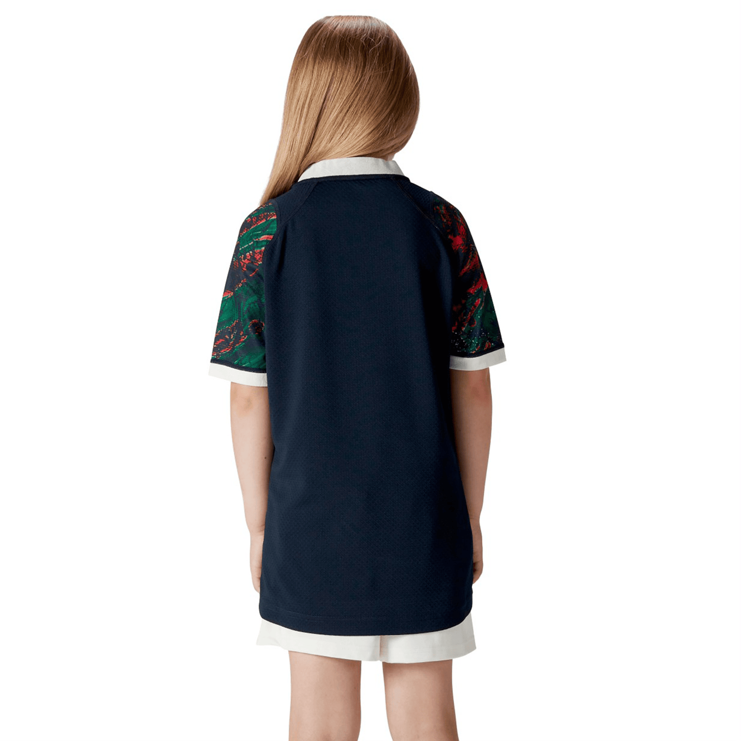 From behind, a child with flowing hair is seen wearing the Youth British & Irish Lions 2025 Warm Up Jersey by Canterbury, featuring a navy-blue color, short sleeves, crisp white collar, and patterned details.