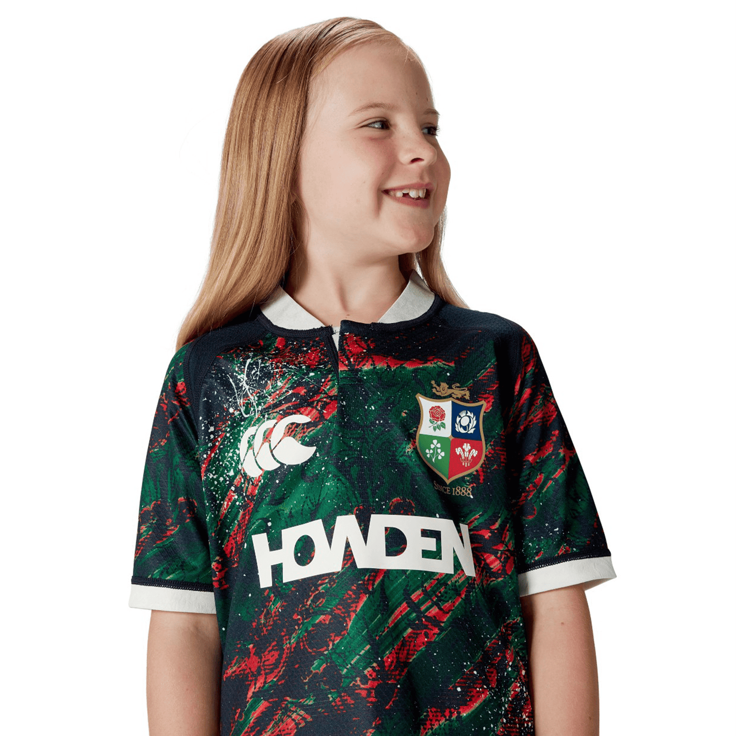 A child smiles while wearing the Youth British & Irish Lions 2025 Warm Up Jersey by Canterbury, glancing to the side against a plain white backdrop. The jersey's moisture-wicking fabric ensures comfort, making it ideal for young Lions fans.