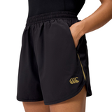 A person wears Canterbury Women's Tempo+ 5" Training Shorts in black with a gold logo on the left thigh, standing with one hand in the pocket. These rugby shorts offer moisture-wicking fabric for all-day comfort.