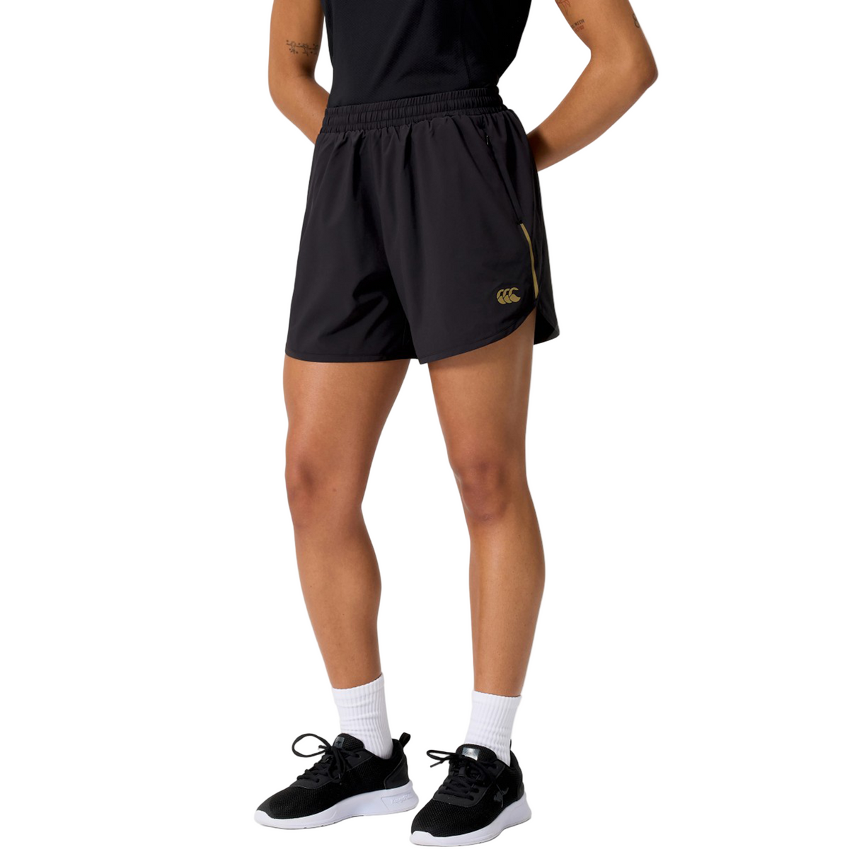 A person wearing a black t-shirt, Canterbury Women's Tempo+ 5" Training Shorts, white socks, and black sneakers stands against a white background.