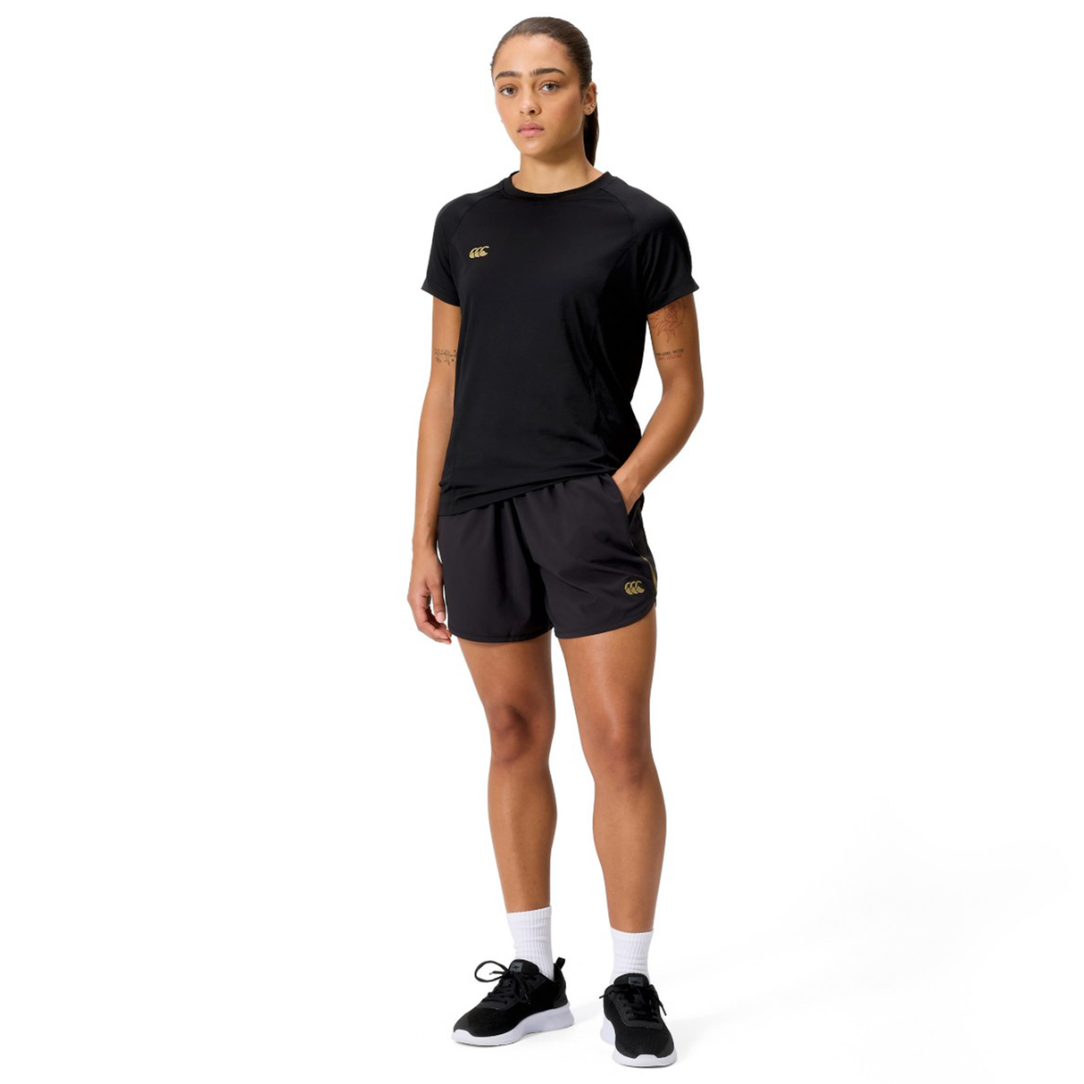 A person faces forward wearing a black t-shirt, Canterbury Women's Tempo+ 5" Training Shorts in black, white socks, and black sneakers against a plain white background.