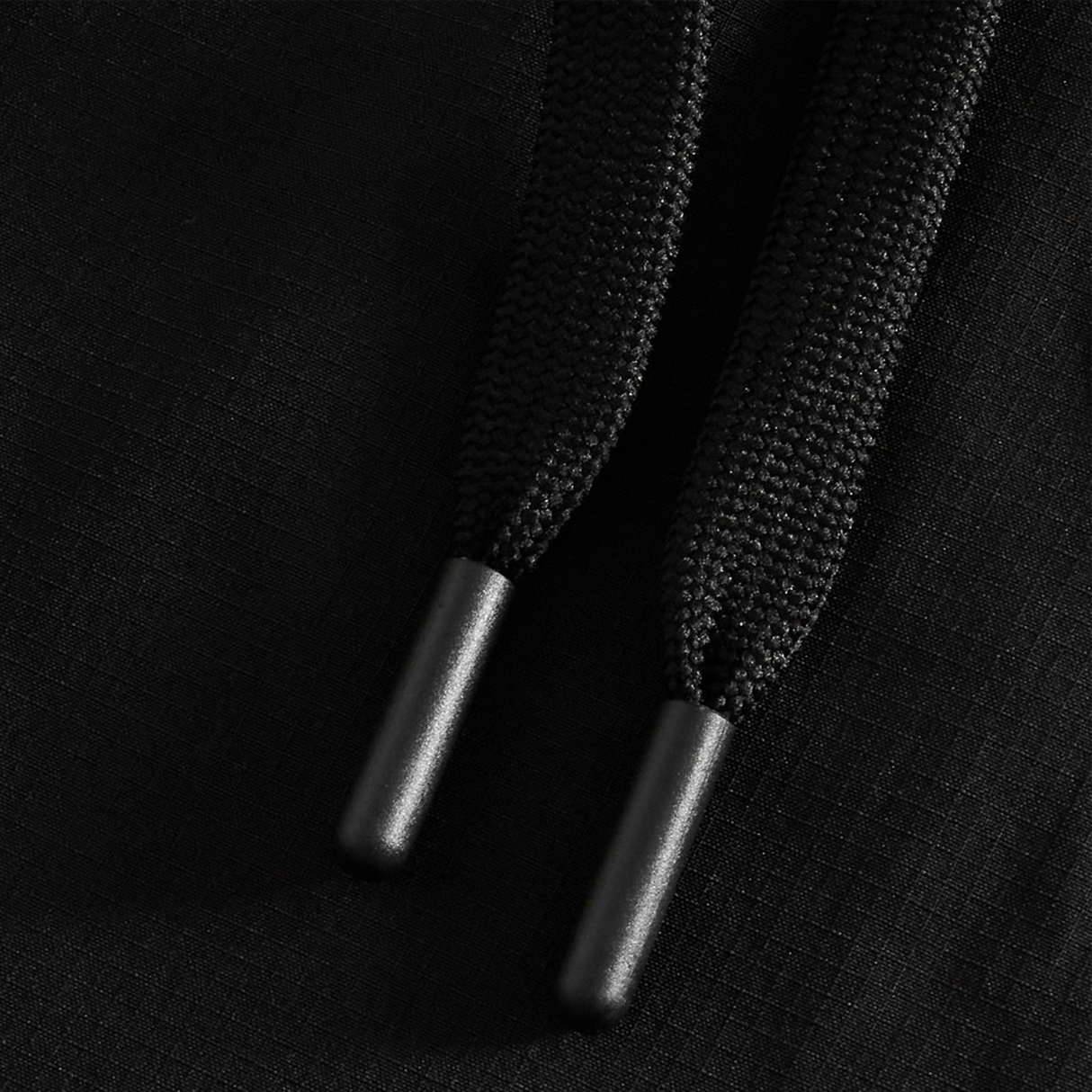 Close-up of two black drawstrings with plastic tips on a black textured fabric background, featured on the Tempo+ 32" Trackpant by Canterbury.