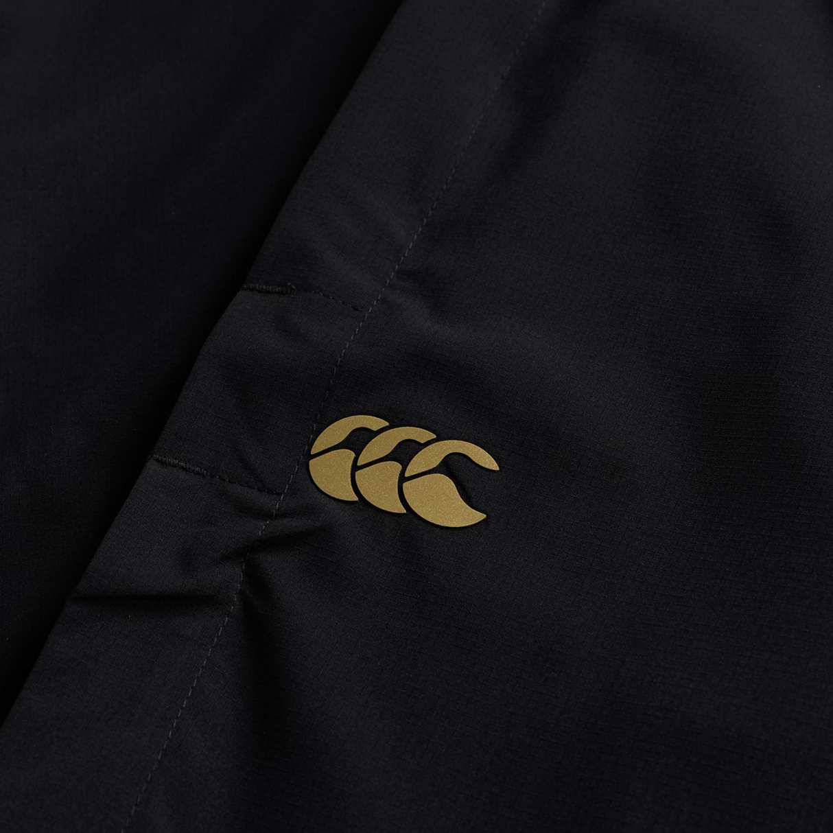 Close-up of the black Tempo+ 32" Trackpant by Canterbury, showing a yellow Canterbury logo with three overlapping shapes printed on the fabric.