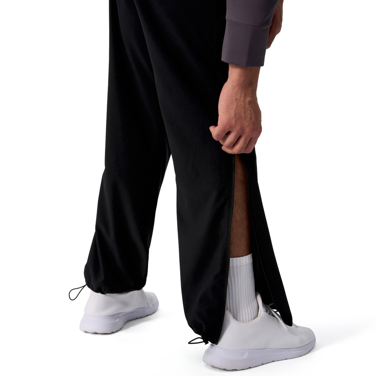 A person wears black Canterbury Tempo+ 32" Trackpant with a partially open side zipper, revealing white socks and white sneakers, standing against a white background.