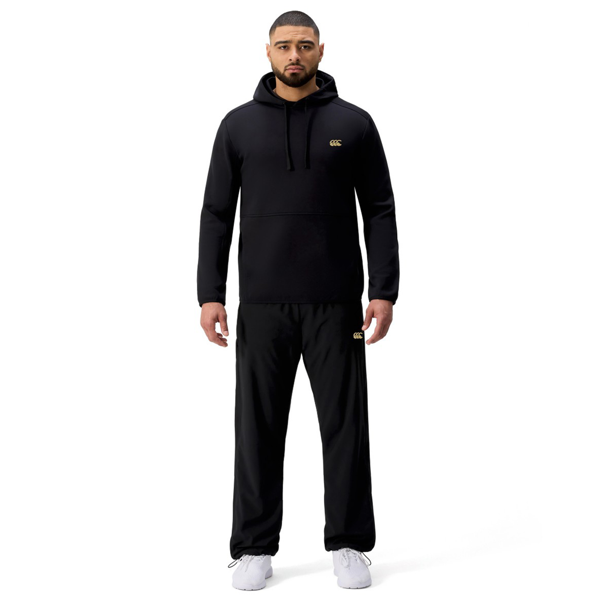 A man stands facing forward wearing the black Tempo+ Hoodie by Canterbury and black pants with white sneakers against a plain white background.