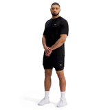 A man stands against a white background wearing Canterbury Tempo+ 5" 2-In-1 Shorts in black, a black athletic t-shirt, white socks, and white sneakers.