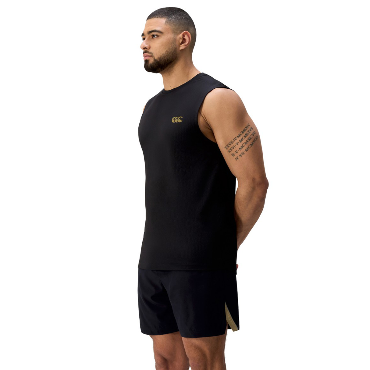 A man in a black Canterbury Tempo+ Training Tank and black shorts stands with his hands behind his back, facing left against a white background.