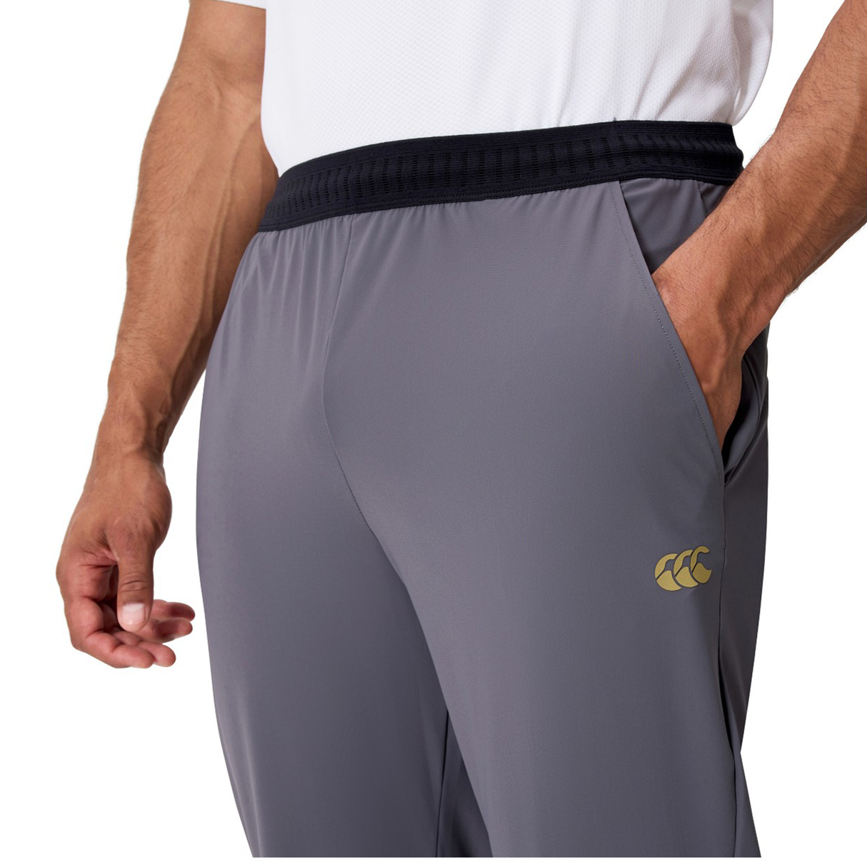 A person wearing a white shirt and Canterbury Everest Woven Pants by Canterbury, featuring a black waistband, yellow logo on the thigh, and one hand in the pocket.