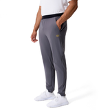 A person stands with one hand in their pocket, wearing a white shirt, white sneakers, and gray Canterbury Everest Woven Pants by Canterbury featuring a small yellow logo.