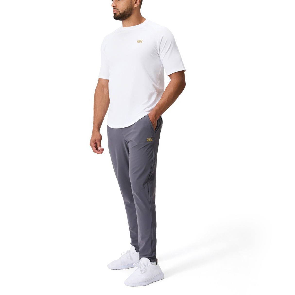 A man in a white short-sleeve shirt, Canterbury Everest Woven Pants by Canterbury, and white sneakers stands with one hand in his pocket against a plain white background.