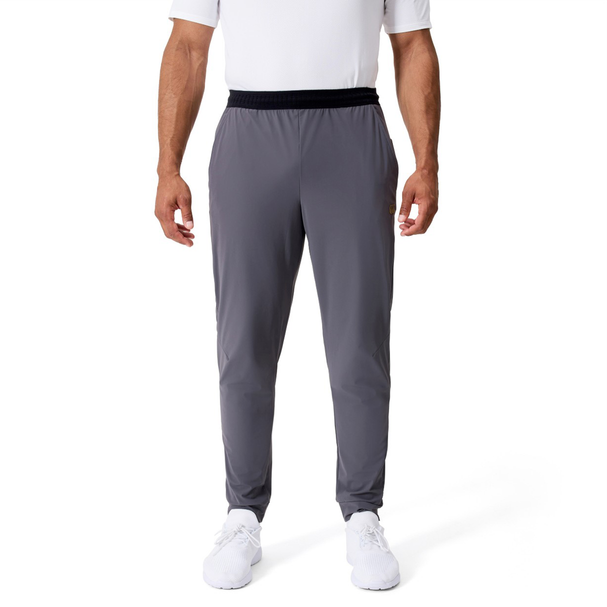 A man stands facing forward against a plain white background wearing a white short-sleeve shirt, Canterbury Everest Woven Pants by Canterbury in gray, and white sneakers.