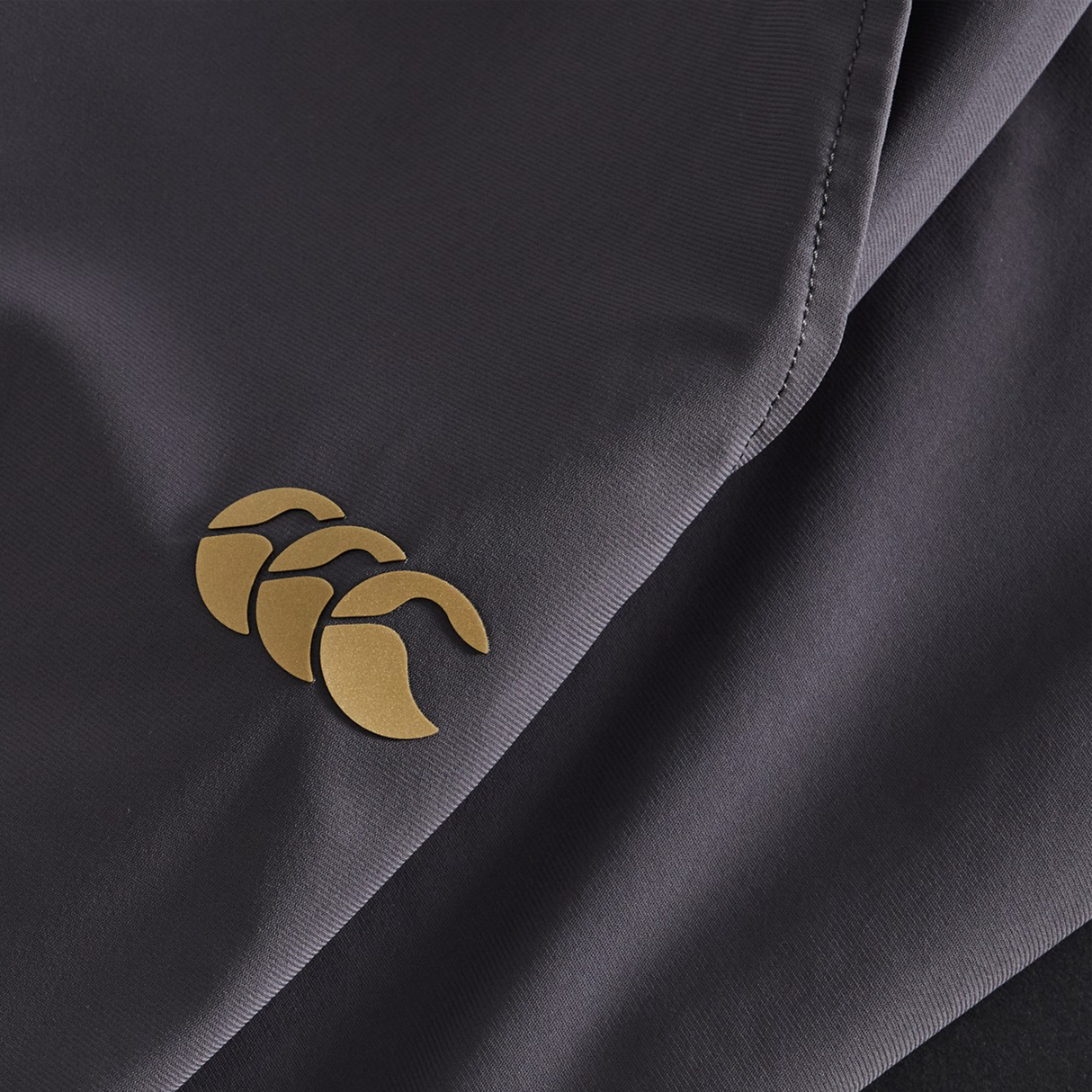 Close-up of Canterbury Everest Woven Pants in dark gray, featuring a gold Canterbury logo on the lower left—ideal for rugby or performance training.
