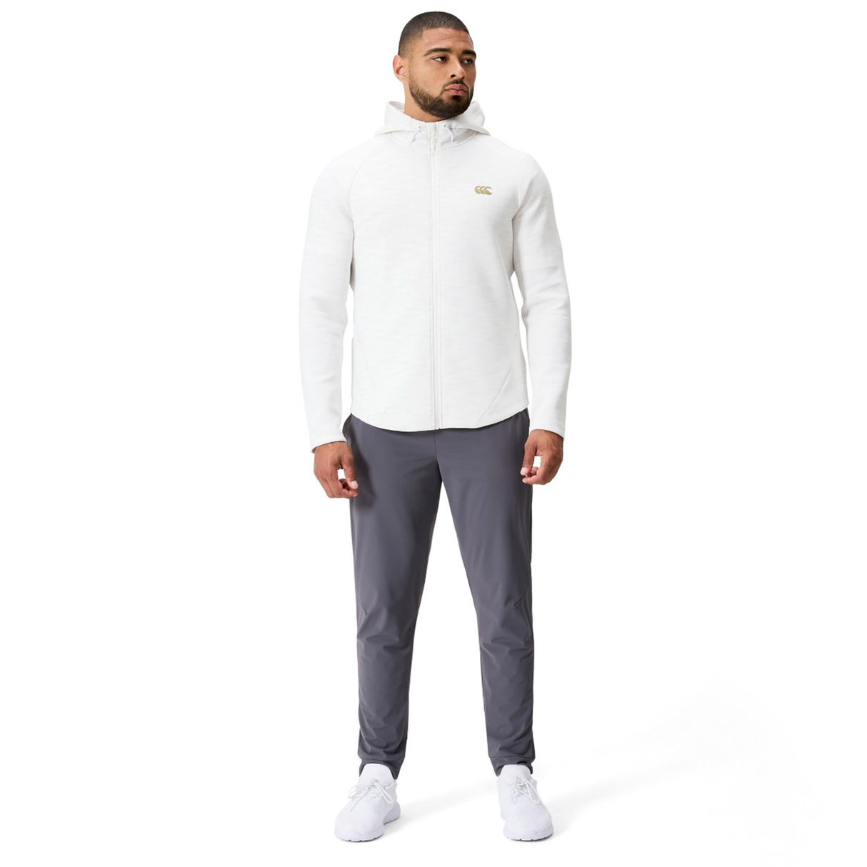 A man faces forward wearing the Canterbury Everest Training Full-Zip Hoodie in white, styled with gray pants and white sneakers, set against a plain white background.